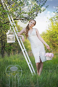 woman wearing white lace dress holding basket leaning on white wooden ladder with birdcage