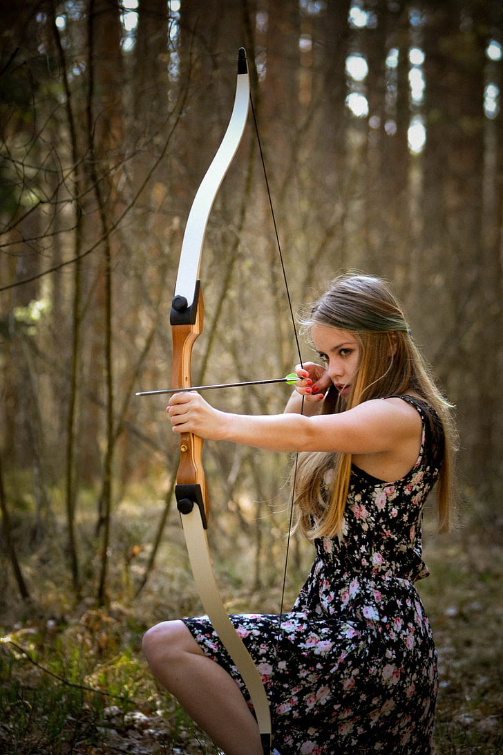 woman in black floral dress holding bow and arrow kneeling in forest