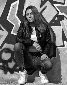 grayscale photography of woman wearing leather jacket and jeans with pair of sneakers