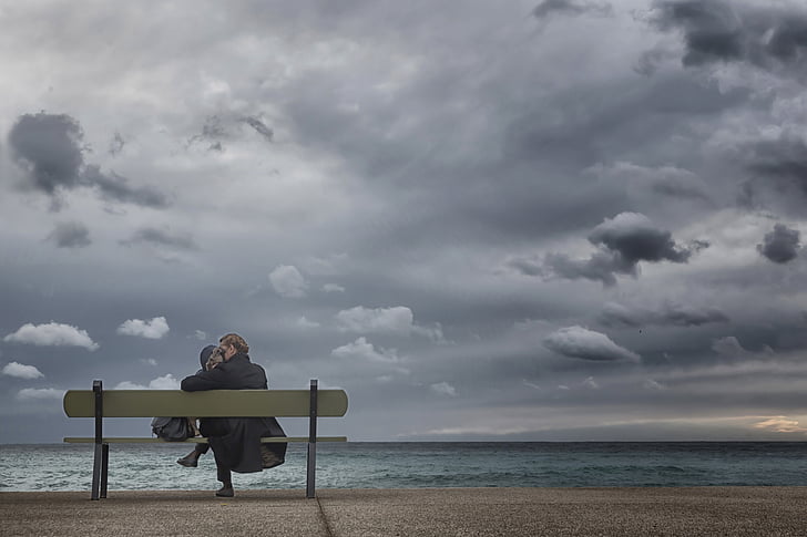 woman sitting on bench facing body of water under cloudy sky