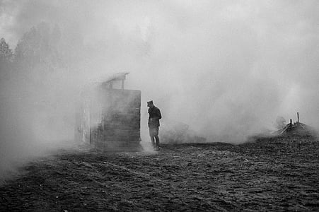 grayscale photography of a man standing next to wooden house with smokes