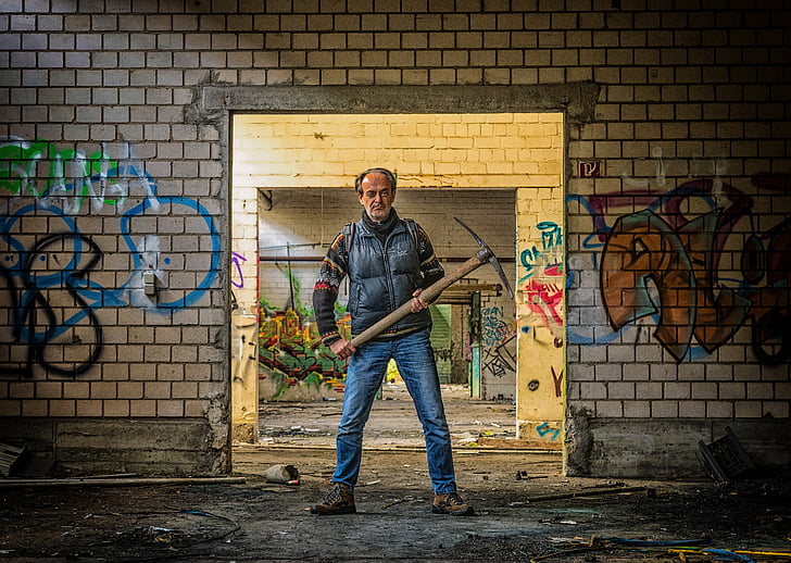 man wearing black vest and blue jeans holding pickaxe behind wall with graffiti