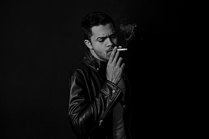 grayscale photography of man wearing black leather jacket holding cigarette stick