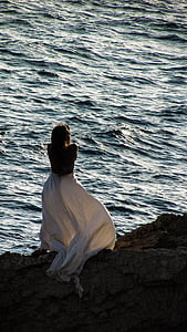 woman wearing white dress facing body of water on brown rock formation