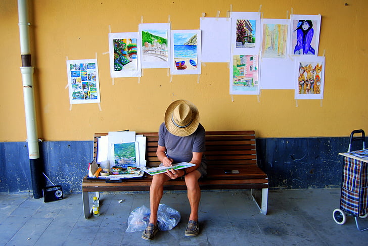 man reading book while sitting on bench near wall pasted with assorted paintings