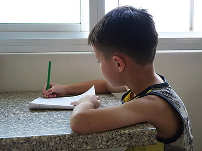 boy wearing white, black, and yellow tank top writing on paper using green pen at daytime