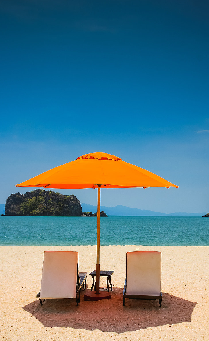 two beach loungers under orange umbrella at seashore under blue calm sky