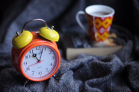 orange and yellow table analog alarm clock near white and multicolored ceramic mug filled with coffee