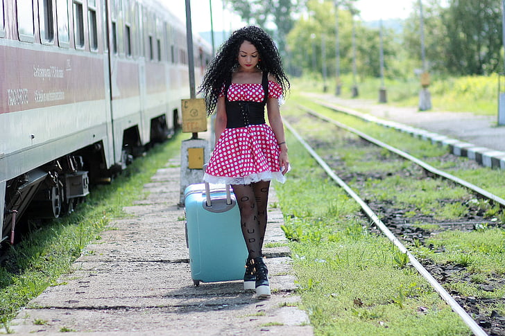 woman in red and white polka-dot dress holding teal luggage beside train during daytime