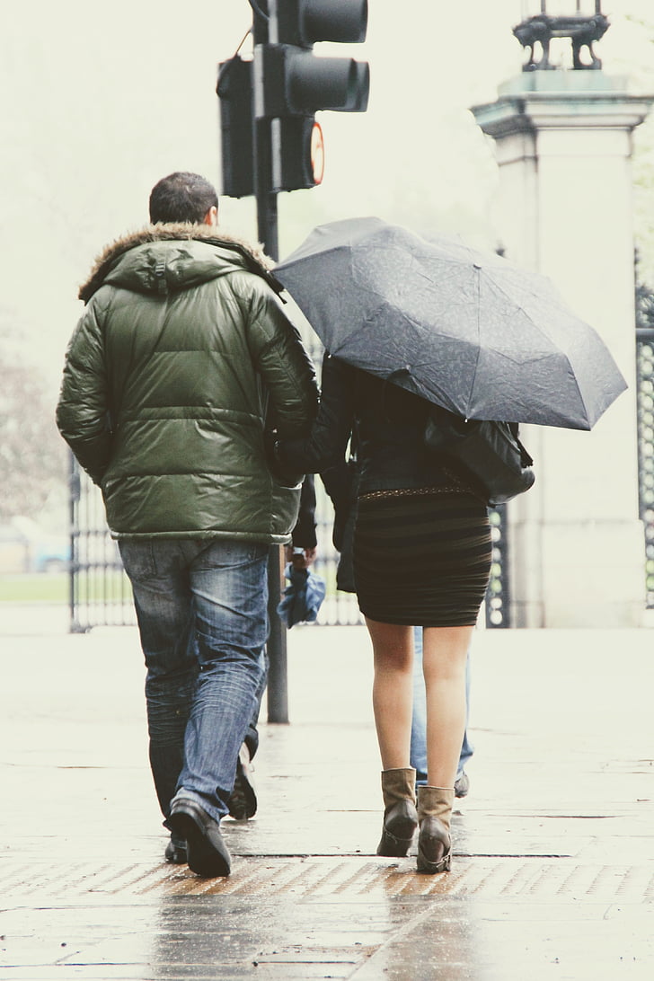 man in green park jacket walking beside woman in black dress with umbrella