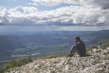 sitting man wearing blue long-sleeved shirt and gray pants in front of green mountain at daytime