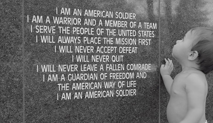 greyscale photo of topless toddler standing in front of I am an American soldier poem-printed wall