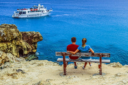 boy and girl seat on brown bench facing on ocean and boat during daytime