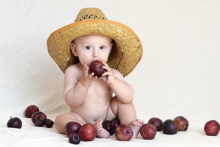 nude baby wearing brown straw hat eating red apple fruit
