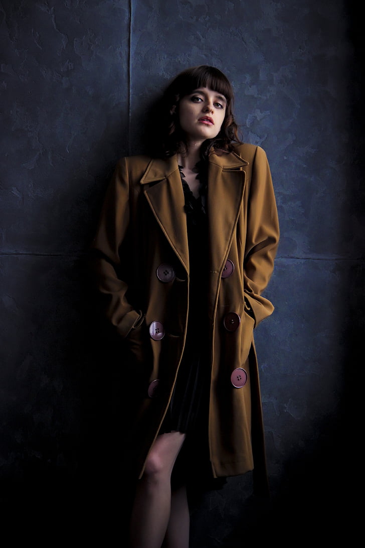 woman wearing brown trench coat stands next to blue painted wall
