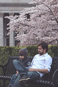 man wearing white button-up long-sleeved shirt sitting on black bench beside toddler wearing gray knitted hat