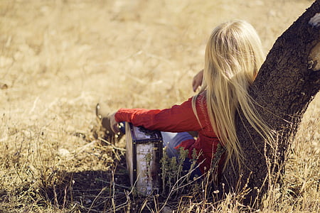 girl with blonde hair wearing red sweater and denim skinny pants leaning on brown tree on beige withered grass field during daytime