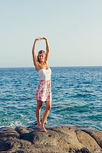 woman stands on rock formation raising her two hands beside body of water