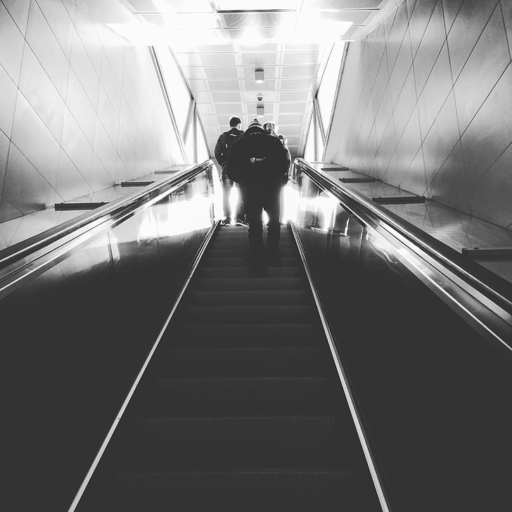 grayscale photo of people climbing using escalator stairs