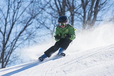 person wearing green ski jacket and snow goggles skiing over snow ground with distance at bare trees during daytime