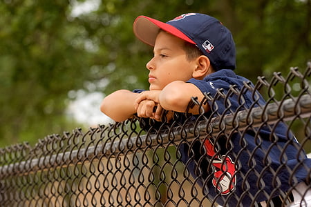 boy wearing blue MLB cap leaning on gray fence