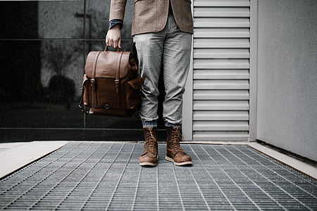 person holding brown leather backpack near gray and black wall