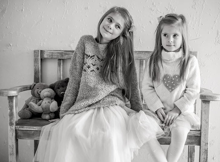 grayscale photo of two girls sits on bench chair with teddy bears