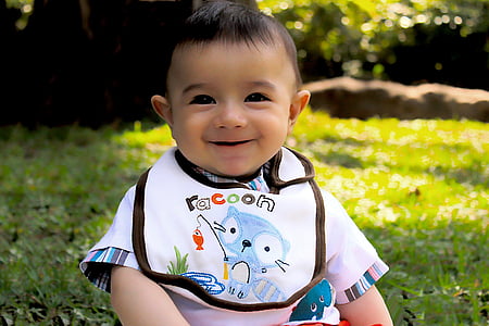 baby in white and multicolored shirt and Racoon bib sitting on green grass at daytime