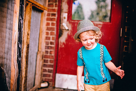 toddler wearing blue crew-neck t-shirt and brown bottoms with gray and black fedora hat near red wooden framed clear glass door during daytime