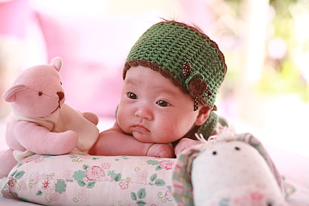 baby wearing green and brown knitted cap near pink teddy bear