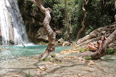 woman wearing black bikini lying on brown tree trunk beside waterfalls during daytime