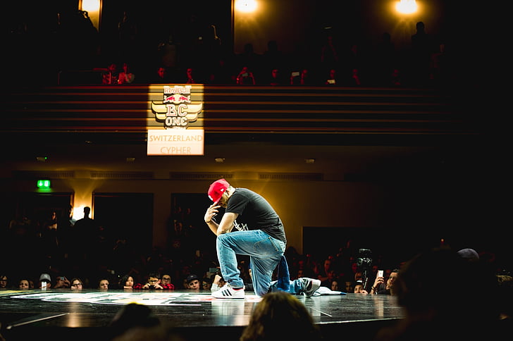 man wearing black t-shirt, red cap and blue denim jeans posing on stage in front of people