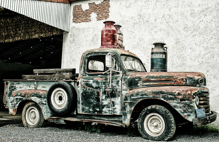 black and brown rusted classic pickup truck park near gray wall with thee assorted-color milk churns on top photo taken during daytime