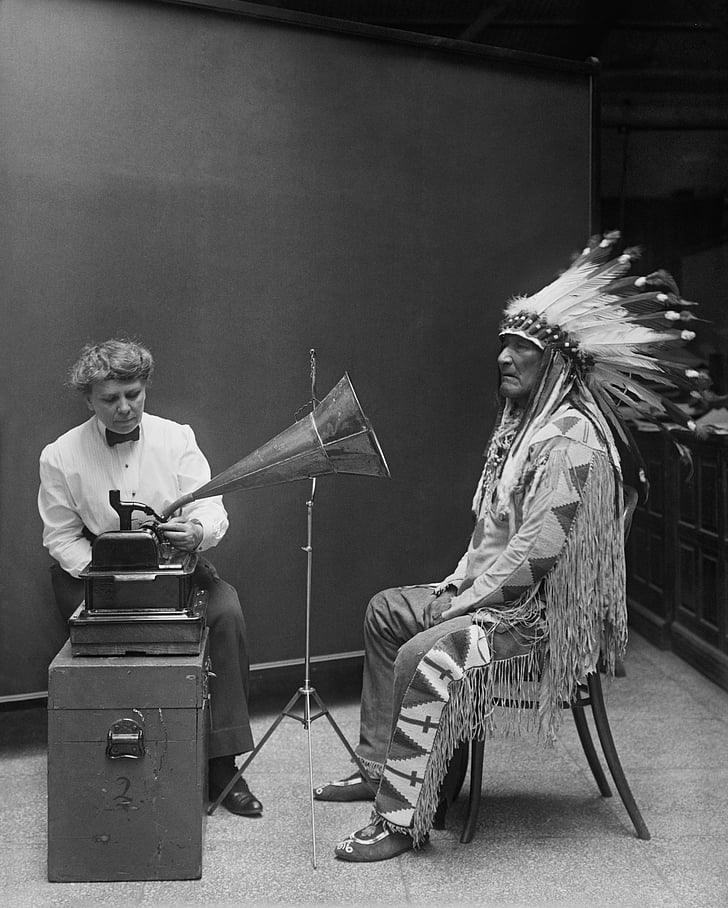 greyscale photo of native American sitting in front of gramophone