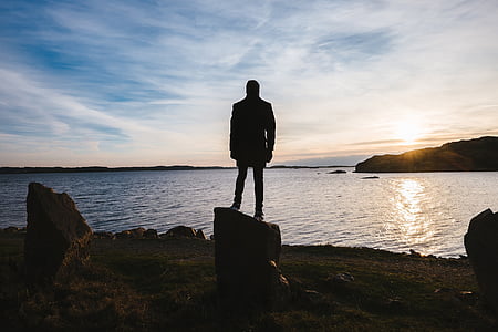 silhouette photo of person standing on rock near body of water during daytime