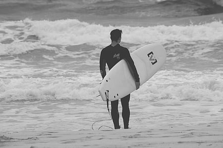 grayscale photography of man carrying surfboard on seashore