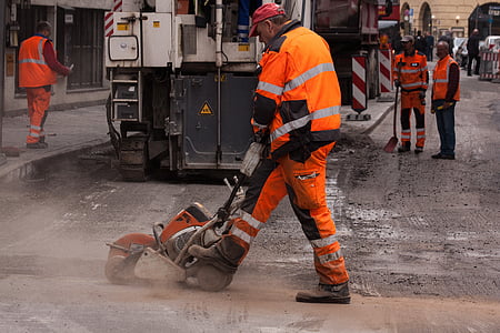man wearing orange and gray neon jacket and pants using concrete saw on road