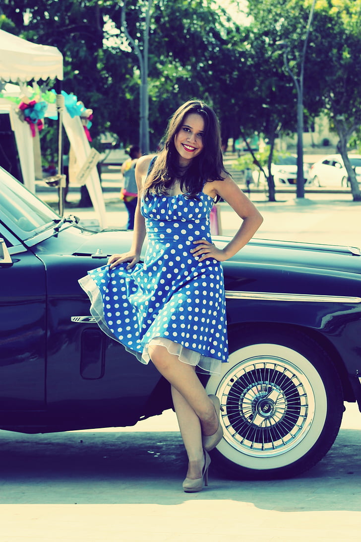 woman in blue and white polka-dot sleeveless dress leaning on black car