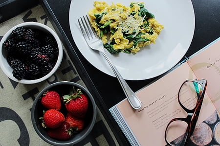 two bowls of blackberries and strawberries next to plate with dish and fork beside opened book with eyeglasses on top