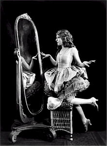 grayscale photography of woman in white sleeveless dress staring her self in vanity mirror