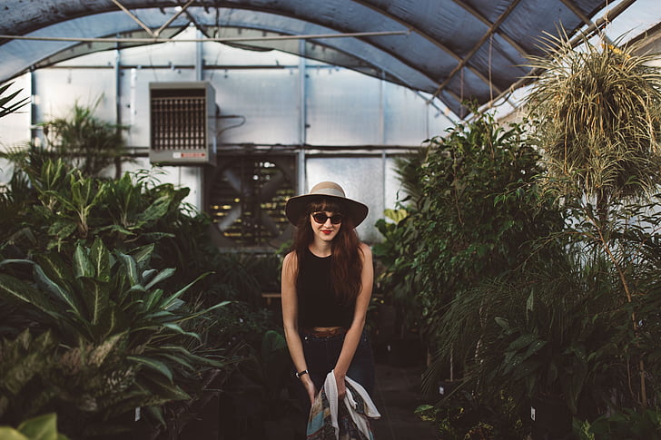 fashion photography of woman in black sleeveless top and gray sun hat standing inside greenhouse