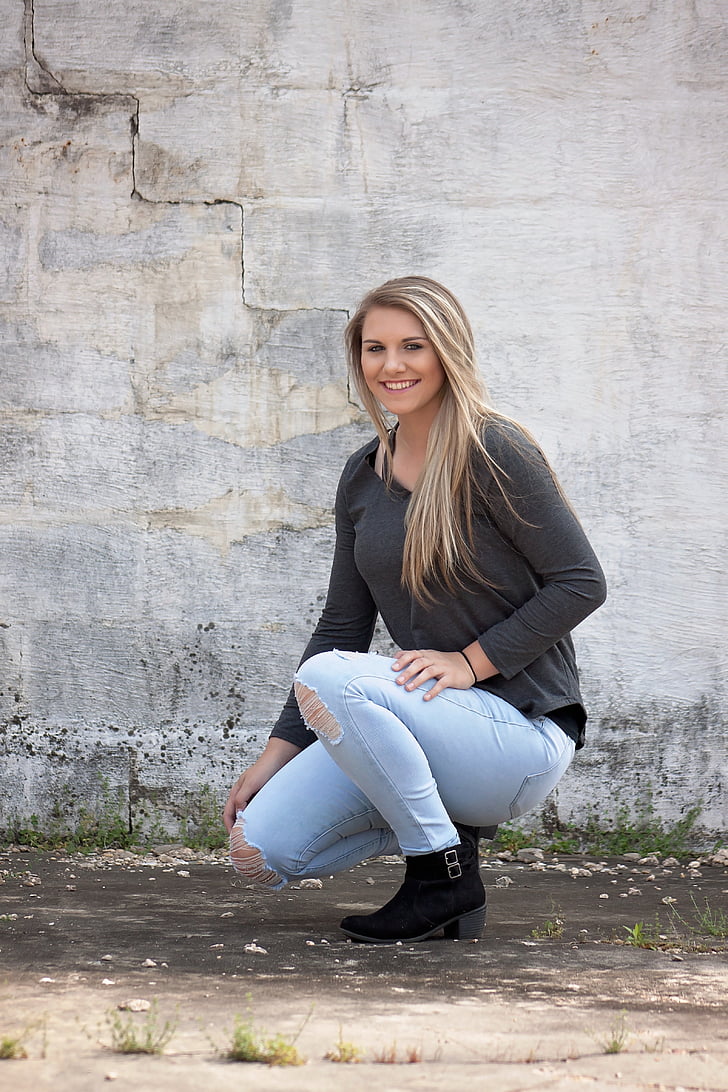 woman wearing grey long-sleeved shirt and distressed blue denim skinny jeans and black buckled boots kneeling near grey concrete wall