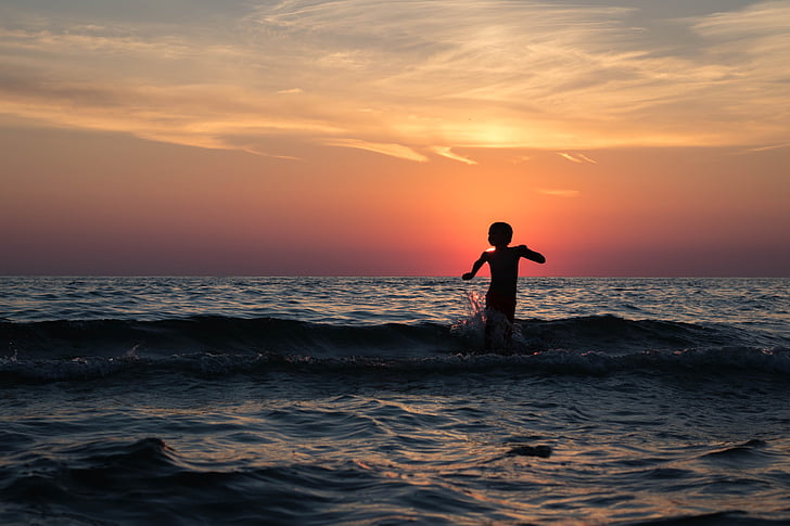child on body of water under yellow and orange sunset