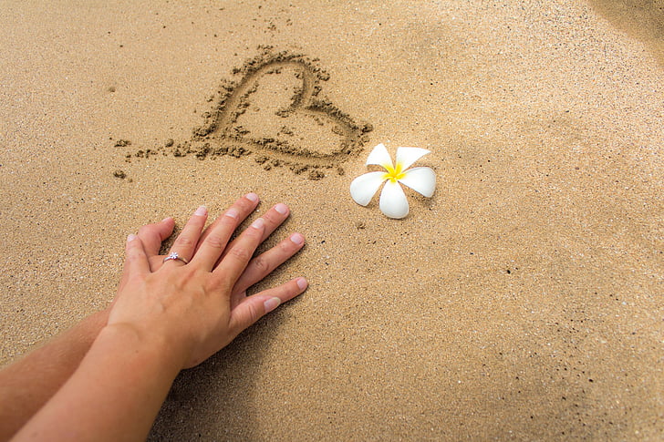 man and woman's hand on white sand with carved heart beside white and yellow plumeria flower