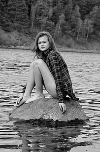 grayscale photo of woman sitting on rock in the middle of body of water