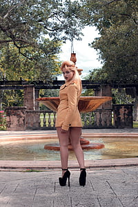 woman wearing brown coat standing beside water fountain near trees under blue sky at daytime