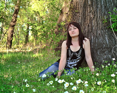 woman wearing black tank top and blue jeans sitting on grass near tree