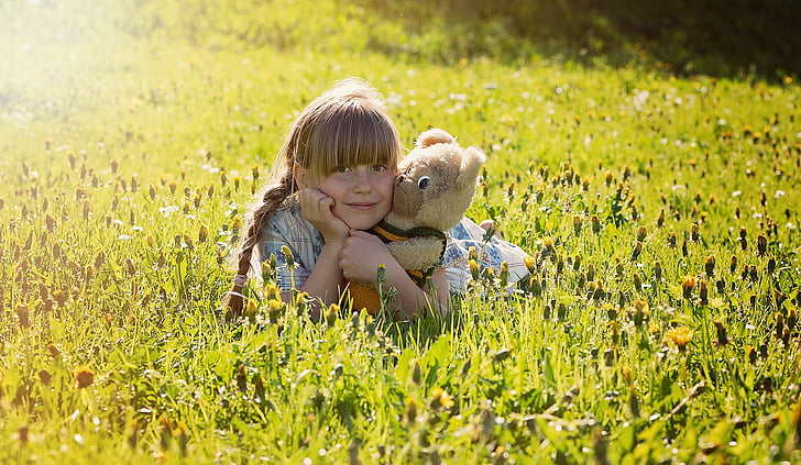 girl lying on green grass field while holding her plush toy