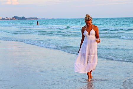 woman in white halter-strap dress walking on seashore during daytime