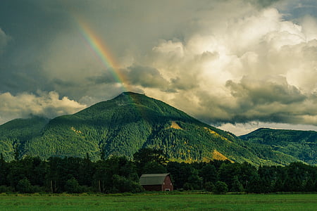 barn near green mountain with rainbow under grey clouds